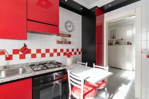 A modern kitchen with red and white cabinetry, a gas stove, a sink, and a dining area with a white table and red chairs. - GuestHost - Comfy Apartment M2 Udine (Milan)