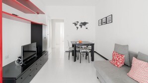 Een moderne woonkamer met een flatscreen televisie, een eethoek met een tafel en stoelen, en een grijze bank met opvallende kussens.