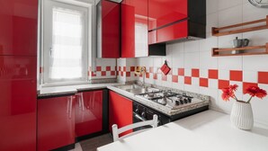 Een keuken met rode kastjes, een gasfornuis, een spoelbak en een raam met zonwering.
