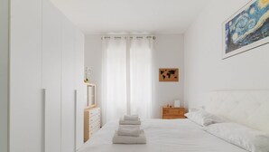 Een slaapkamer met een bed, witte gordijnen, een houten nachtkastje, een wereldkaart in een lijst en een schilderij.