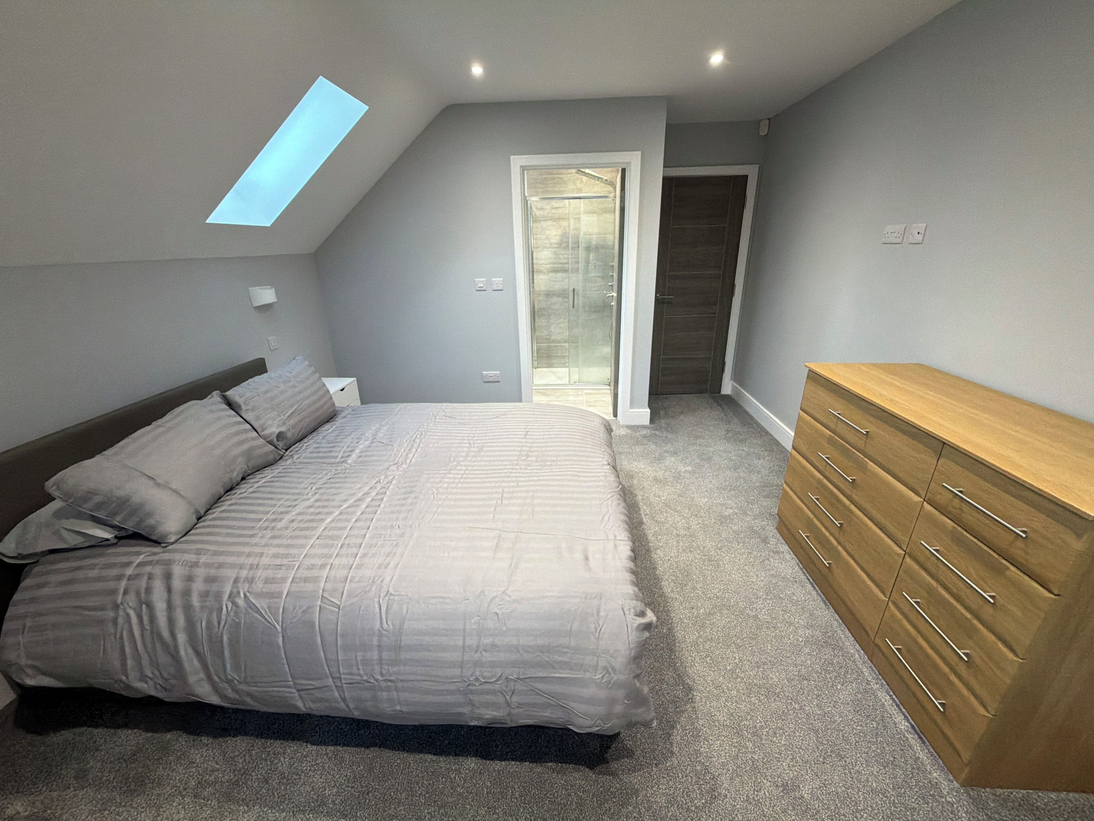 A bedroom with a bed, dresser, skylight, and a door leading to a bathroom.