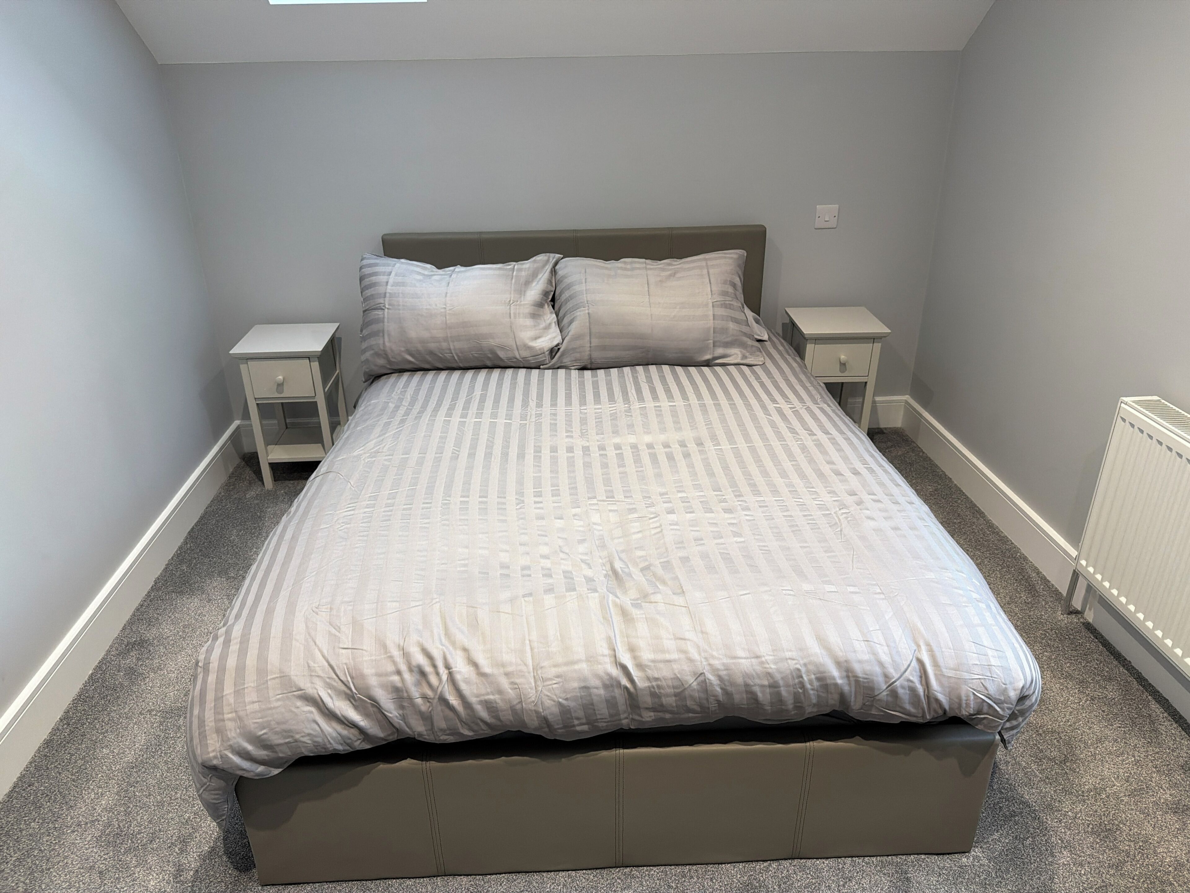 A single bed with a headboard, two bedside tables, a radiator, and a carpeted floor.