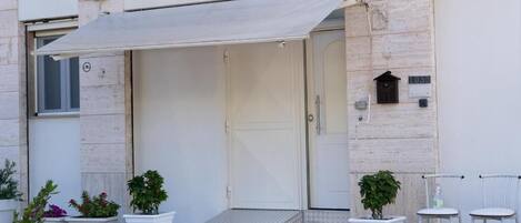 A building entrance with a covered porch, potted plants, and two white chairs.
