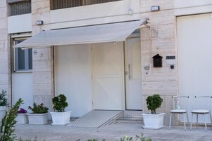 Una entrada de edificio con porche cubierto, plantas en macetas y dos sillas blancas.