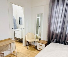 Un dormitorio con cama, escritorio, silla, espejo y baño visible a través de una puerta abierta.