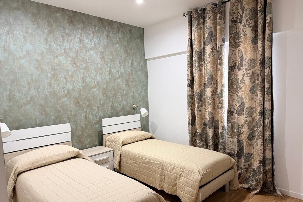 Un dormitorio con dos camas individuales, una mesita de noche con lámpara y un ventanal con cortinas.