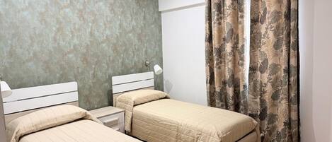 A bedroom with two single beds, a nightstand with a lamp, and a large window with curtains.