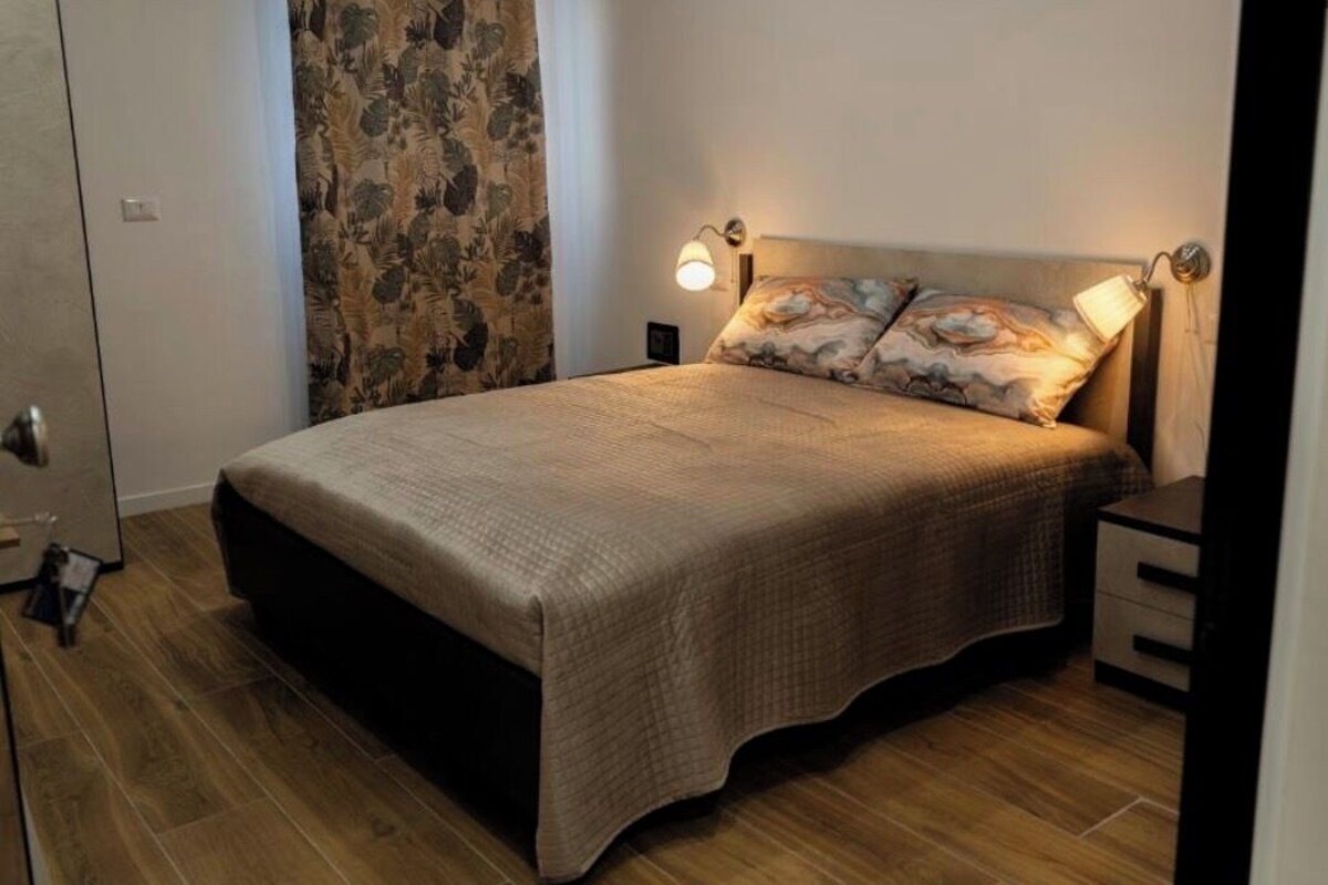 Un dormitorio con cama, mesitas de noche, lámpara y cortina.