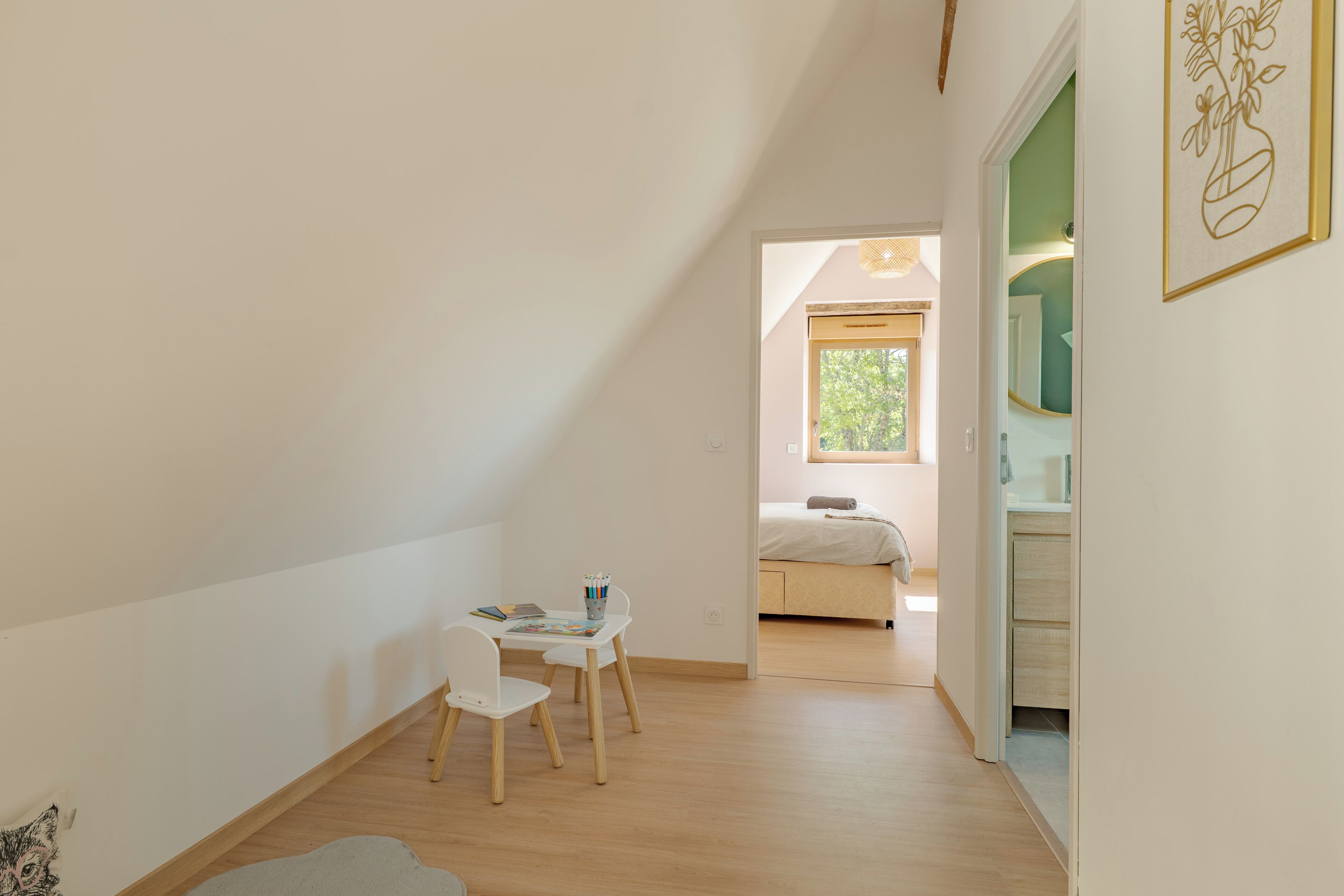 Una habitación con techo inclinado, un escritorio pequeño con sillas, una cama y un baño con espejo.