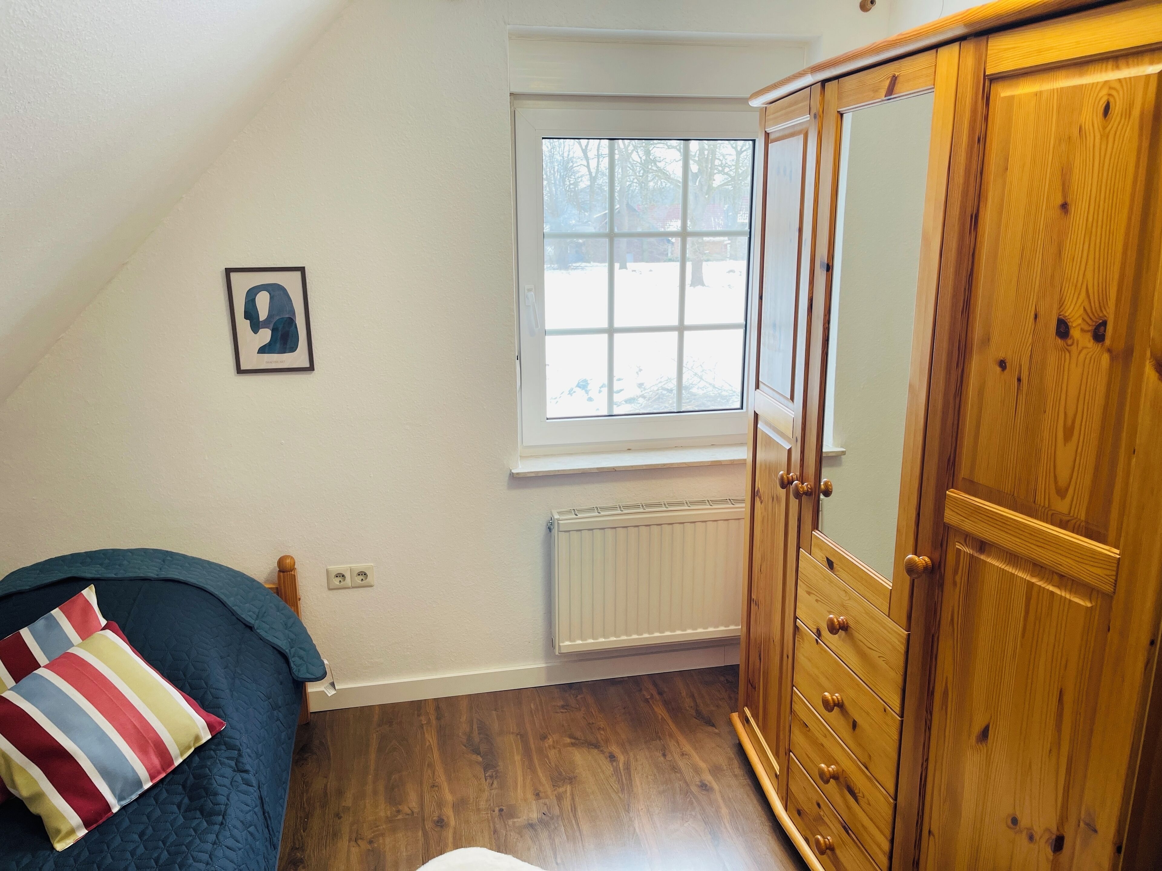 Un dormitorio con techo inclinado, una ventana con vista a árboles, un armario de madera y una cama con cubrecama acolchado.