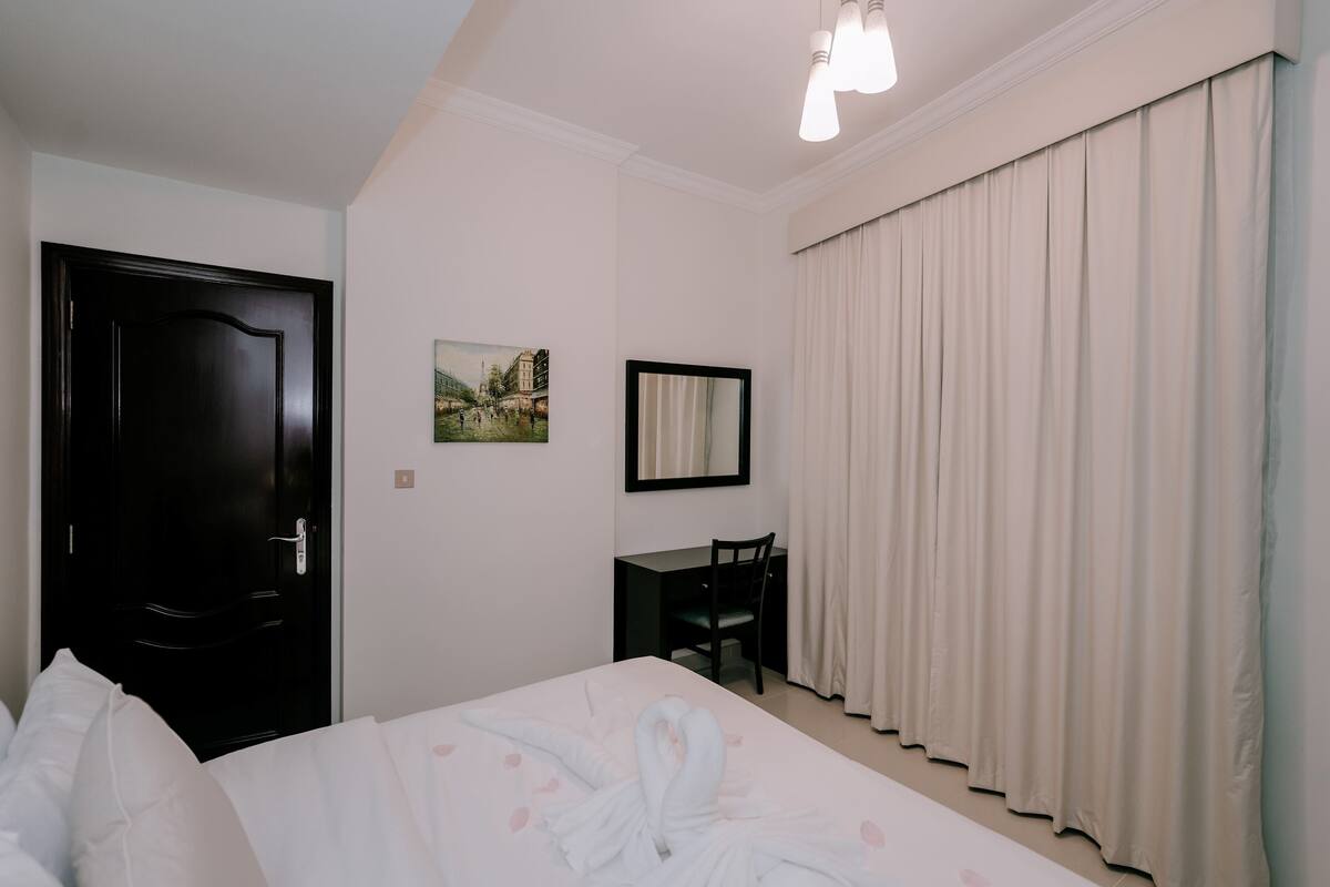 Habitació d'hotel amb un llit, una taula de treball, una cadira, una porta, una finestra amb cortines i un quadre a la paret.