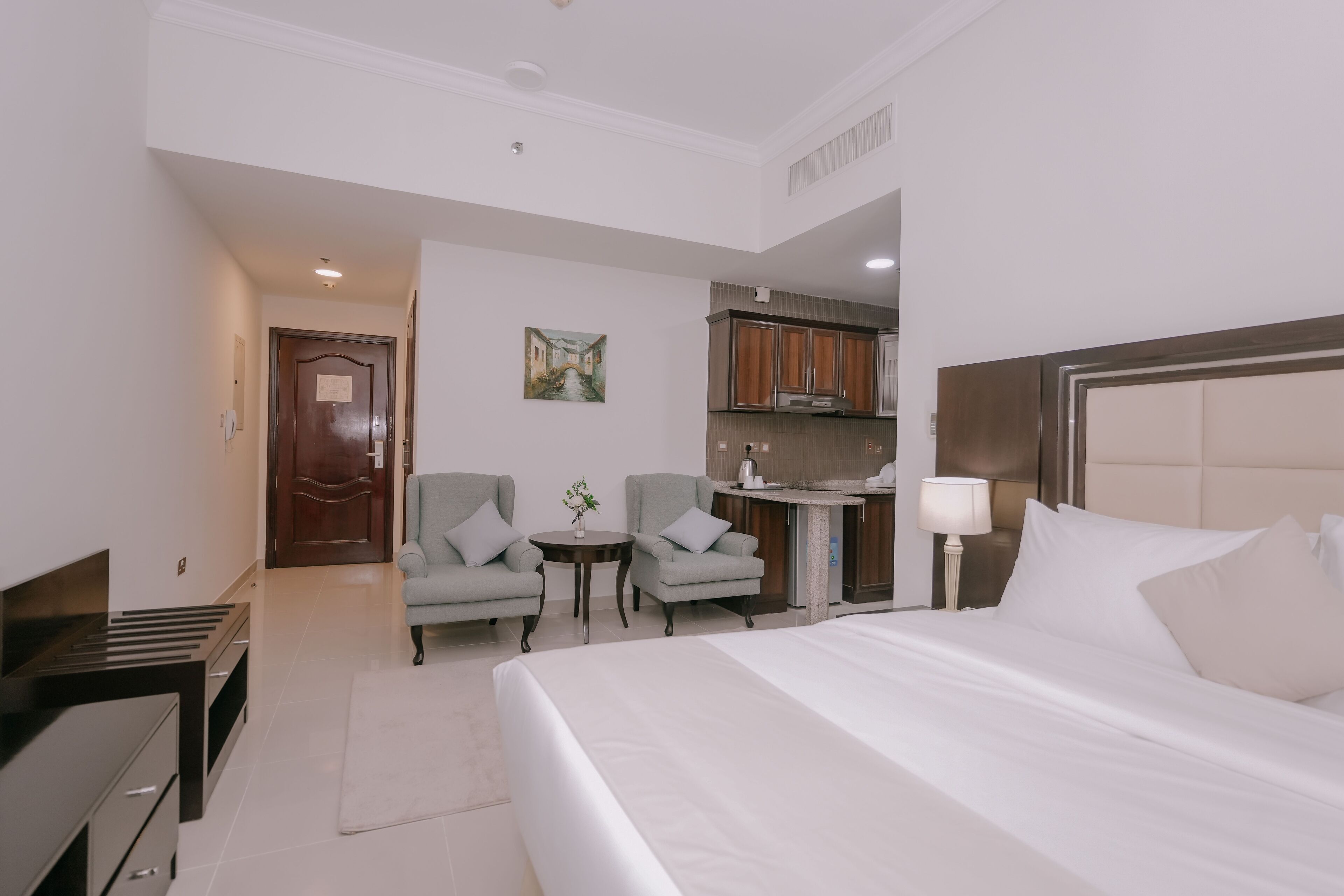 Habitació d'hotel amb un llit, escriptori, dues butaques, una taula petita amb un ram i una cuineta al fons.