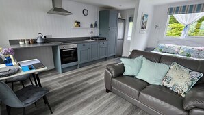 A modern kitchen with grey cabinets, a built-in oven, and a sink. There's a dining area with a table and chairs, and a brown leather sofa with floral cushions. - 1 Bed Cabin-sleeps 2, Hot Tub, Garden, Views, Pets (Callington)