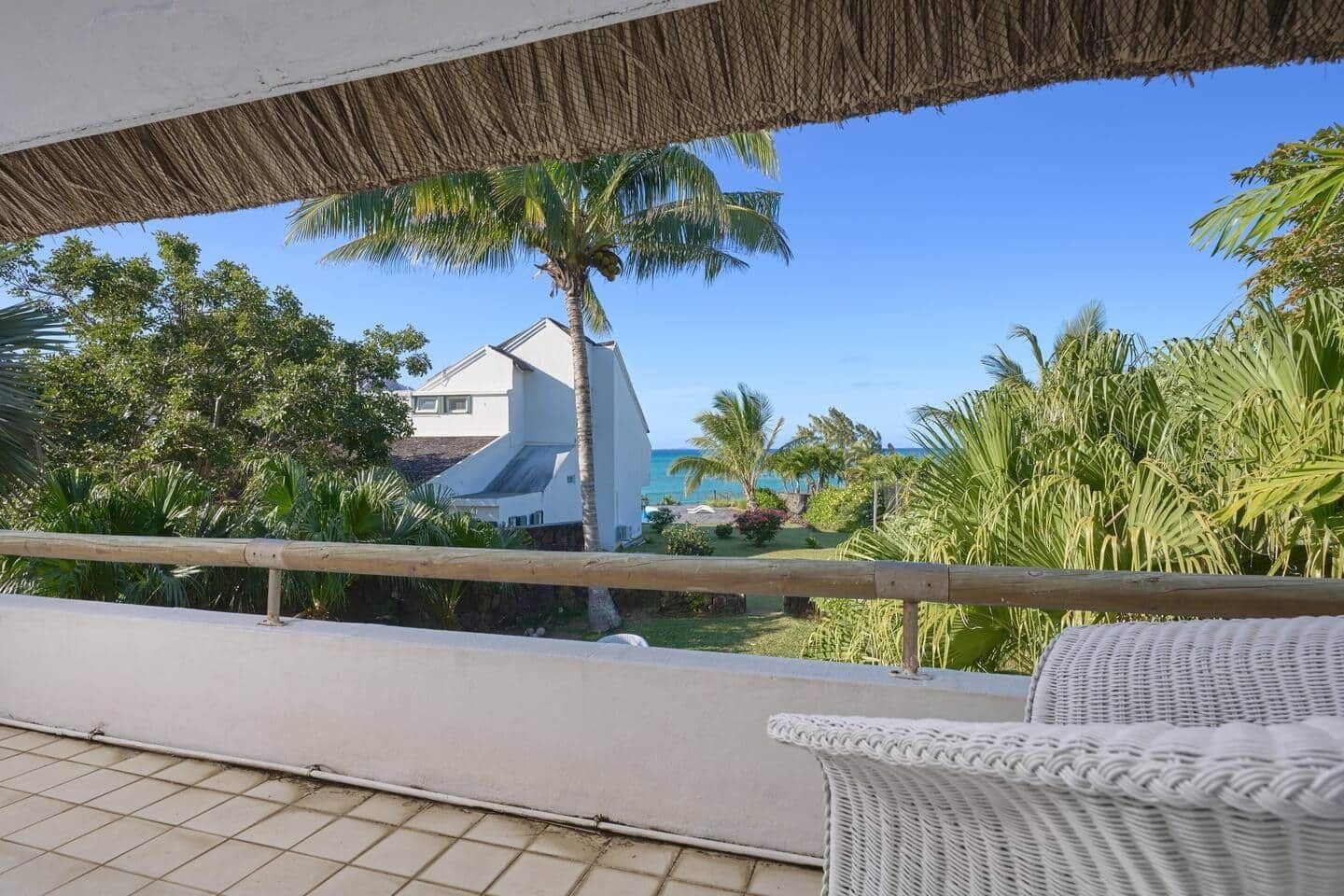 A balcony with a wicker chair, a thatched roof, and a view of a building and palm trees.