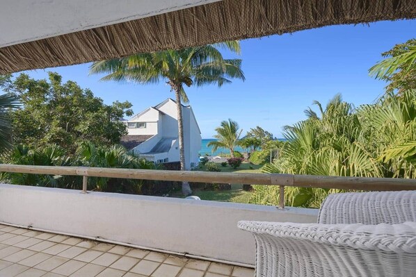 A balcony with a wicker chair, a thatched roof, and a view of a building and palm trees.