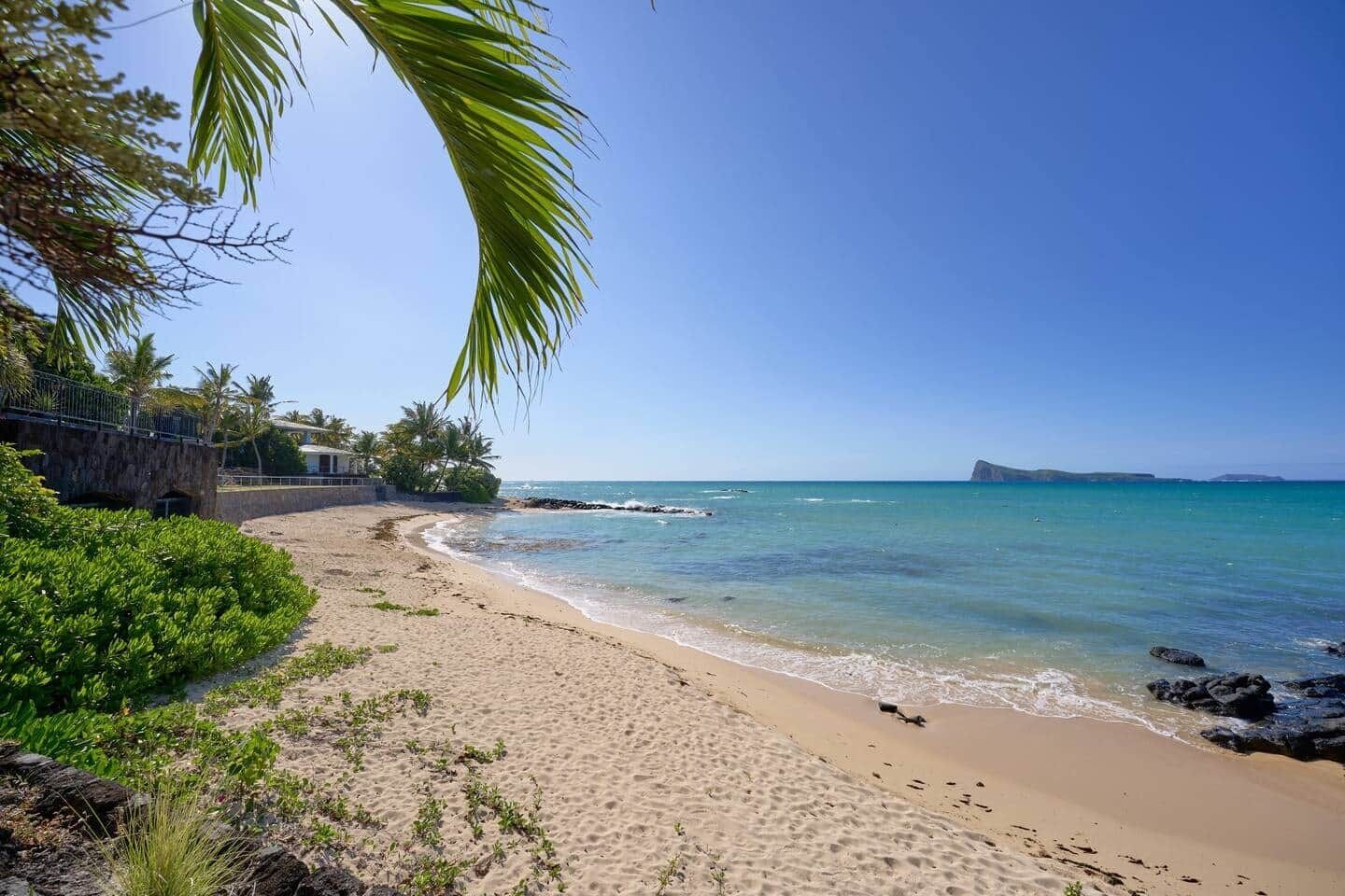 A beach with clear turquoise water, a sandy shore, and a rocky outcrop in the distance.