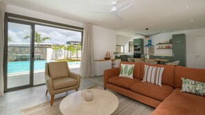 Una sala de estar moderna con un sofá, un sillón y una mesa redonda. Se ve una cocina al fondo y una piscina a través de las puertas de cristal.