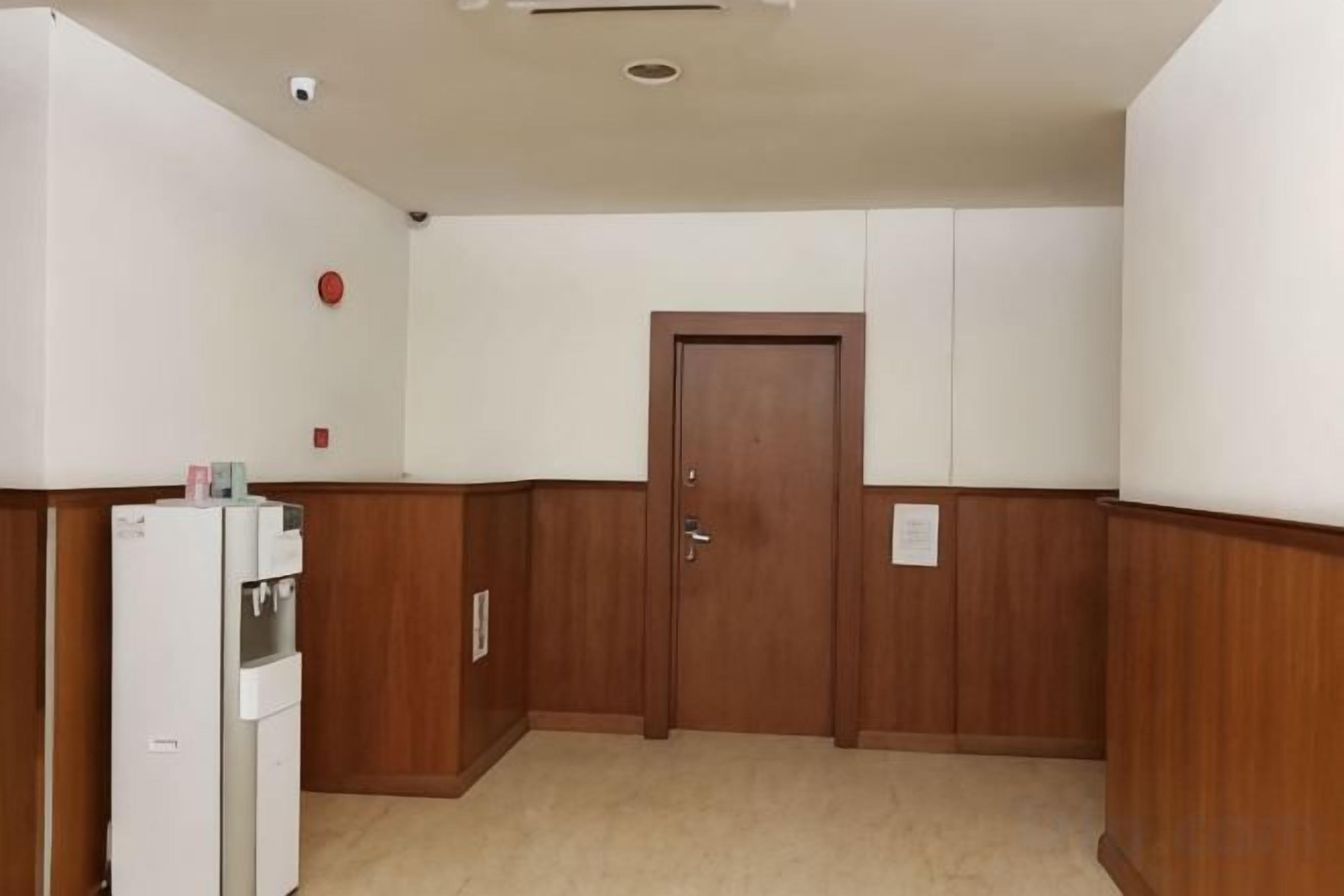 A hallway with a wooden door, a white refrigerator, and a wall-mounted security camera.