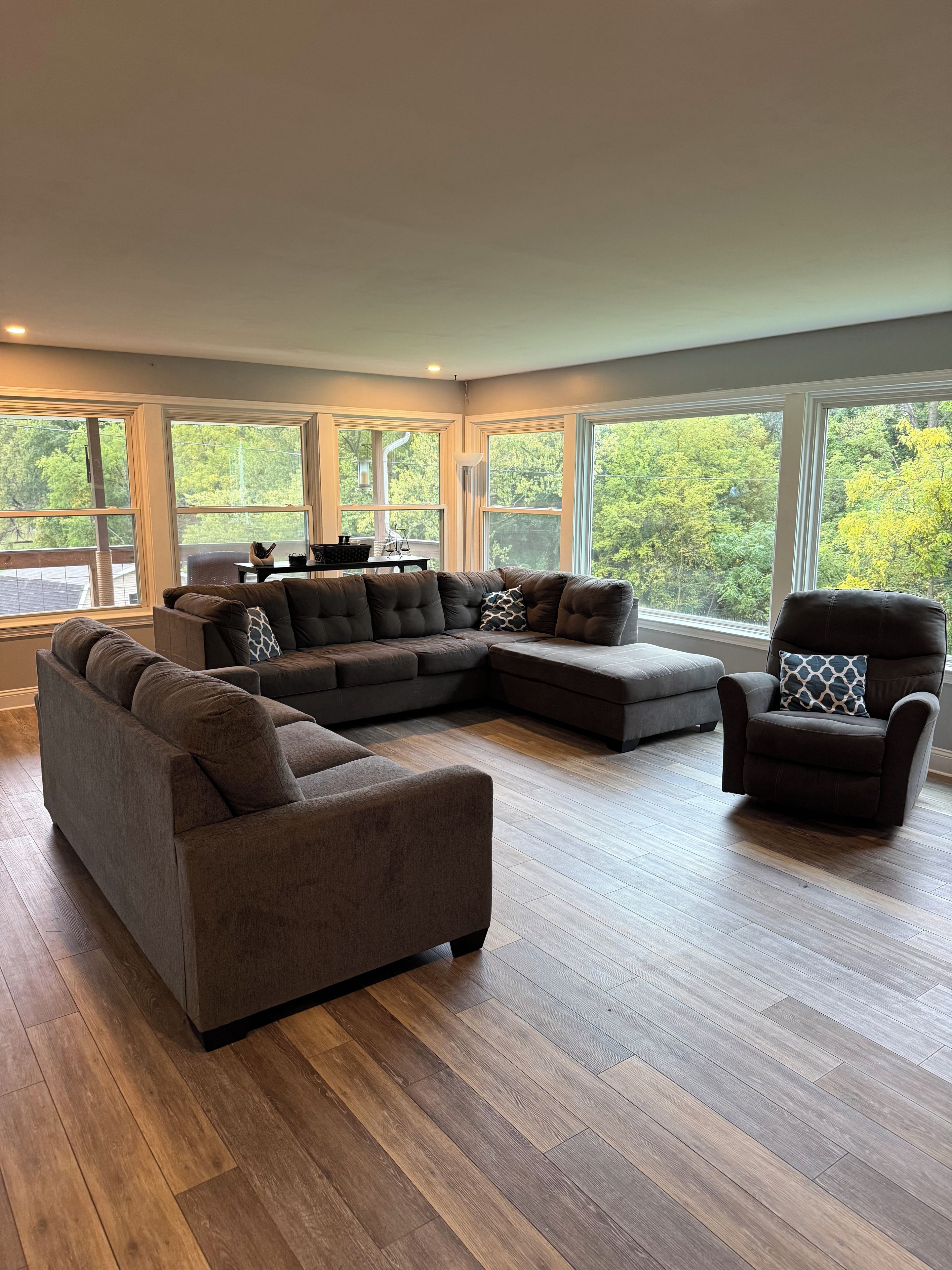 A modern living room with a sectional sofa, a recliner, and a coffee table. Large windows provide a view of the outdoors.