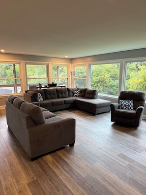 Una sala de estar moderna con un sofá modular, un sillón reclinable y una mesa de centro. Amplios ventanales ofrecen vistas al exterior.