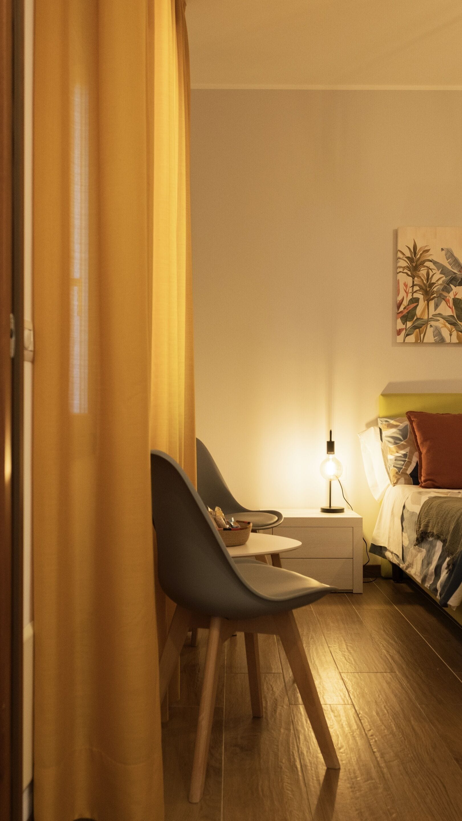 A room with a bed, a bedside table with a lamp, a chair, and a desk with a chair.