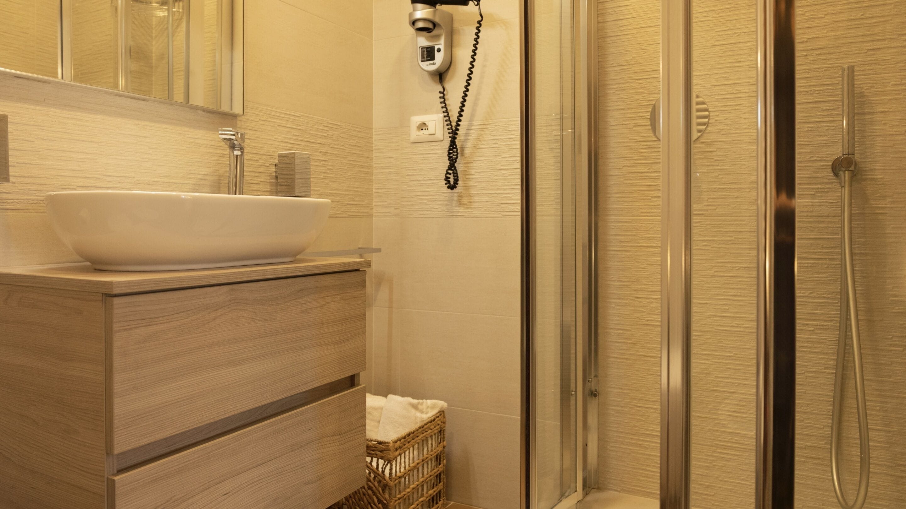 Bagno con lavandino bianco, mobile bagno in legno, uno specchio, doccia con porta in vetro e telefono fissato alla parete.