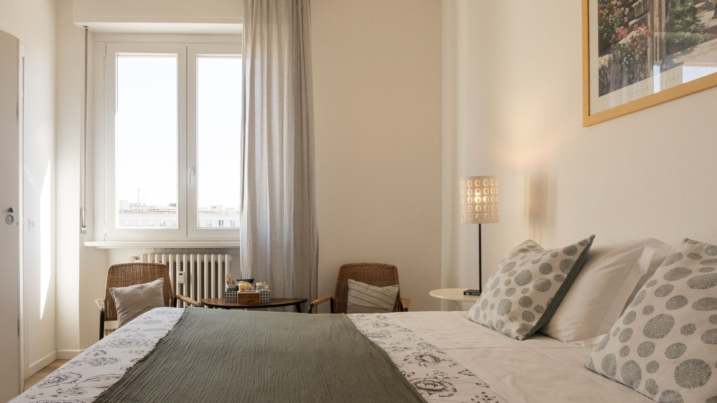 A hotel room with a bed, two chairs, a table, a radiator, and a window with curtains.