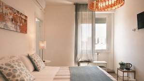 Un dormitorio con cama, almohadas, una mesita de noche con lámpara, una ventana con cortinas y un televisor montado en la pared.