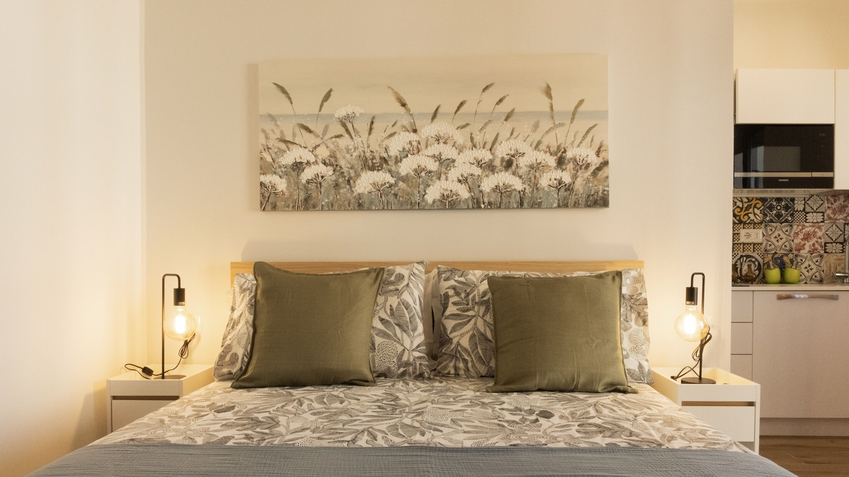 Une chambre avec un lit, deux tables de chevet, un tableau accroché au mur et un coin cuisine.