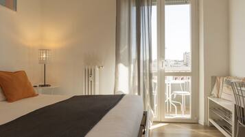 A hotel room with a bed, a desk, a chair, a radiator, and a view of the city through a balcony door.