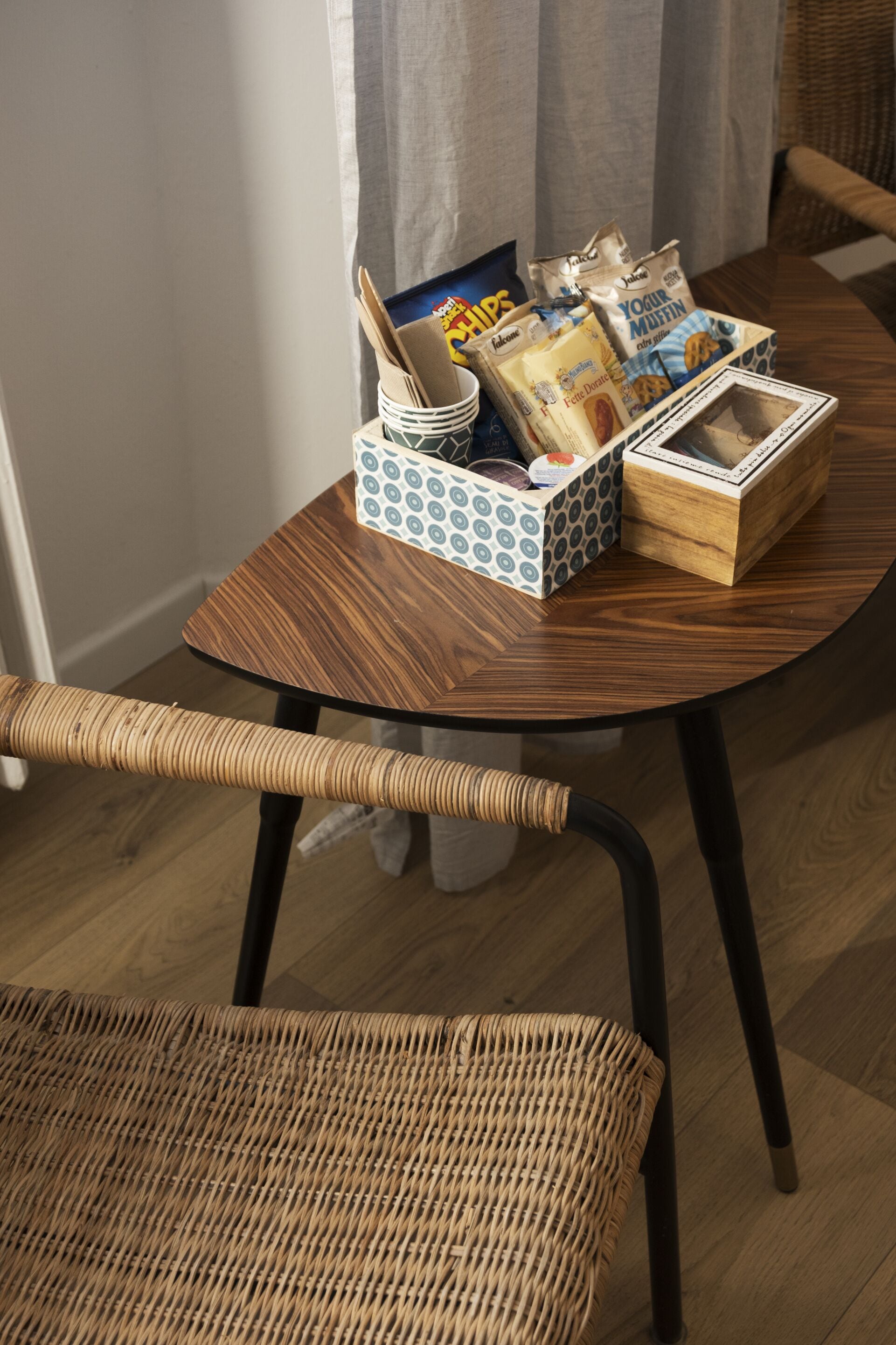 Una mesa redonda de madera con una silla de mimbre, una cesta pequeña con aperitivos y una caja de yogur.