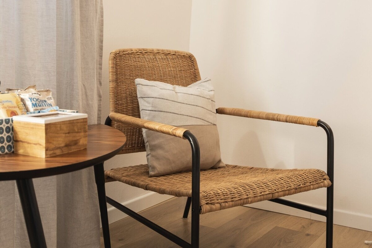 A wicker chair with a cushion, a small round table with a box and bags, and a curtain.