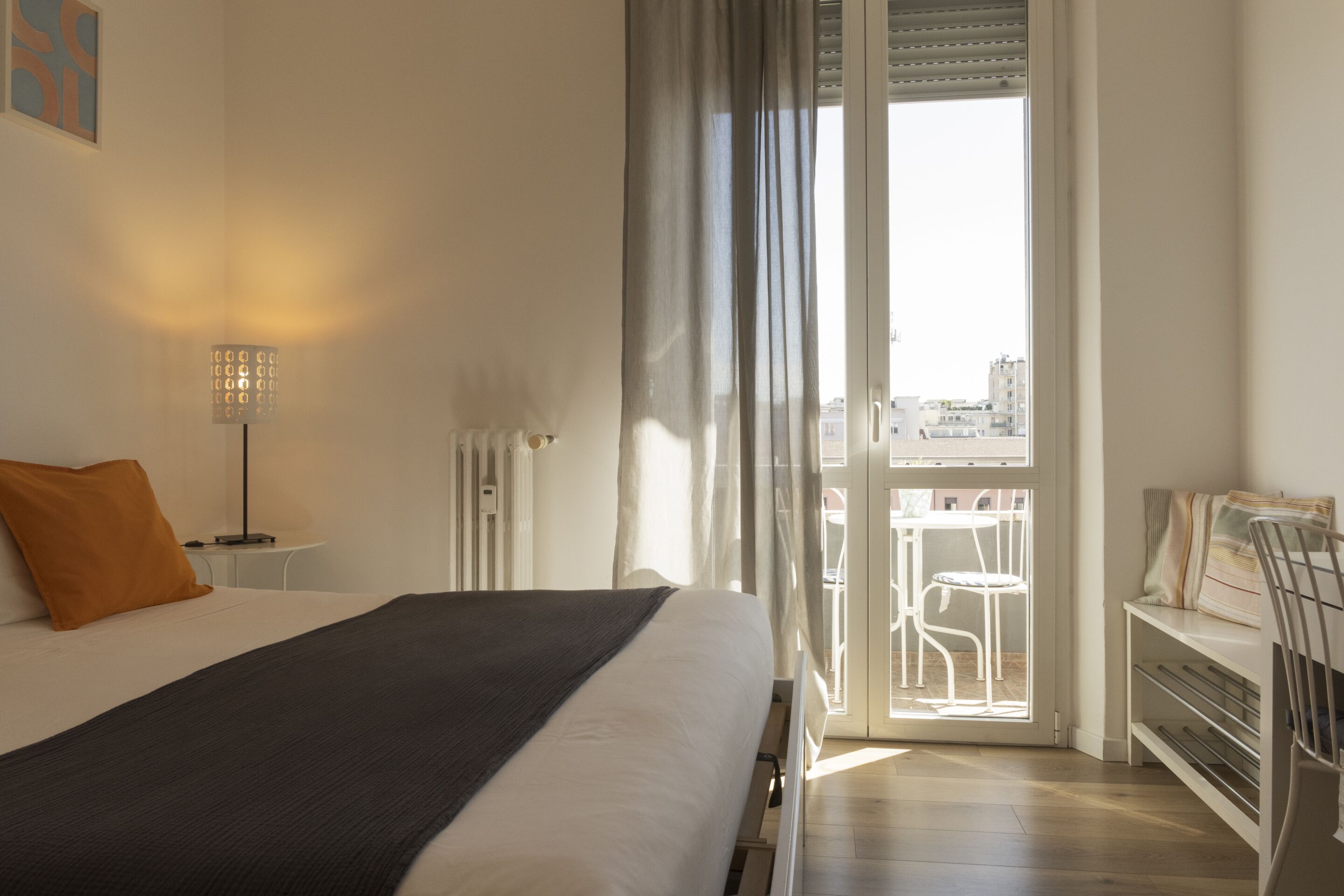Ein Hotelzimmer mit Bett, Schreibtisch, Stuhl, Lampe und Blick auf die Stadt durch ein Fenster.