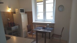 A compact kitchen with a refrigerator, microwave, and a small table with chairs. A clock is visible on the wall, and a window provides natural light. - Perfect for Old Town, Chopin and Warsaw Universities Visits. Safe, quiet, budget (Warszawa)