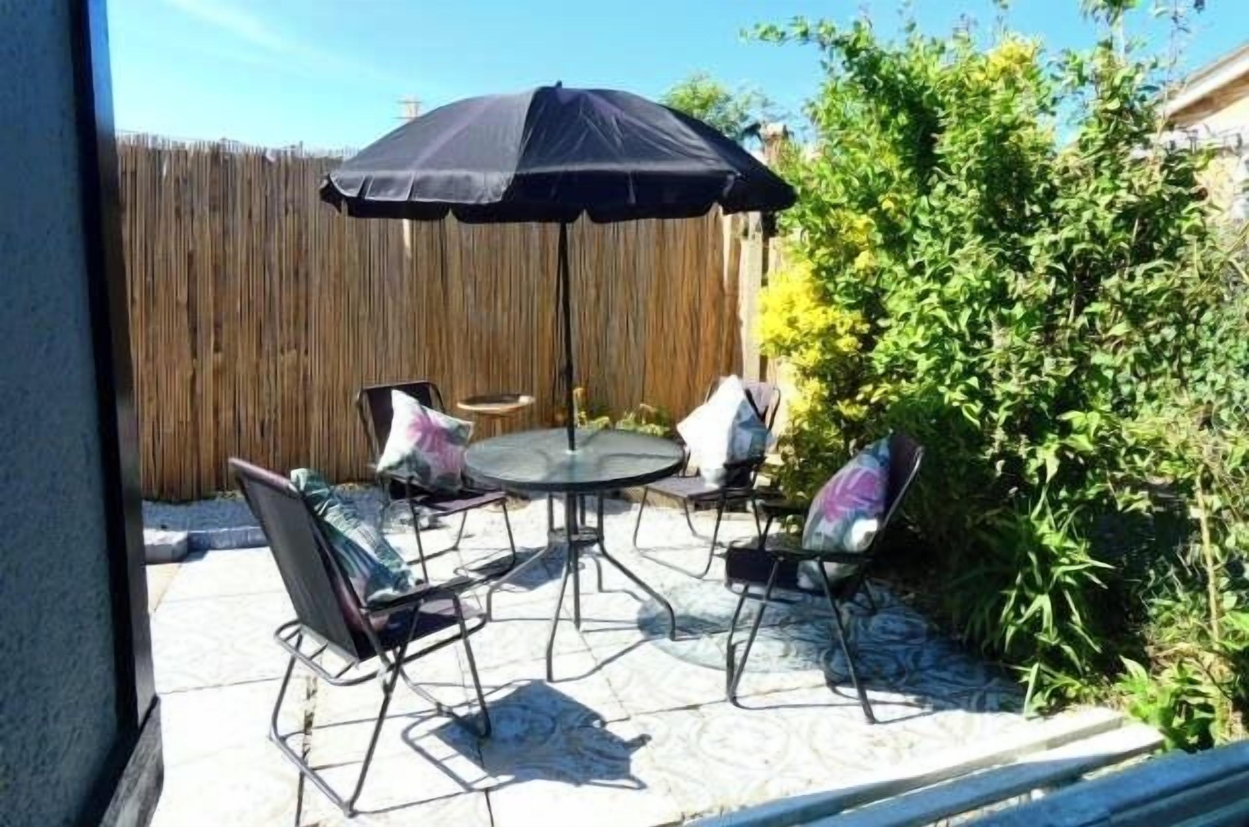 Une terrasse extérieure avec une table ronde et quatre chaises, un parasol noir et une clôture en bois en arrière-plan.