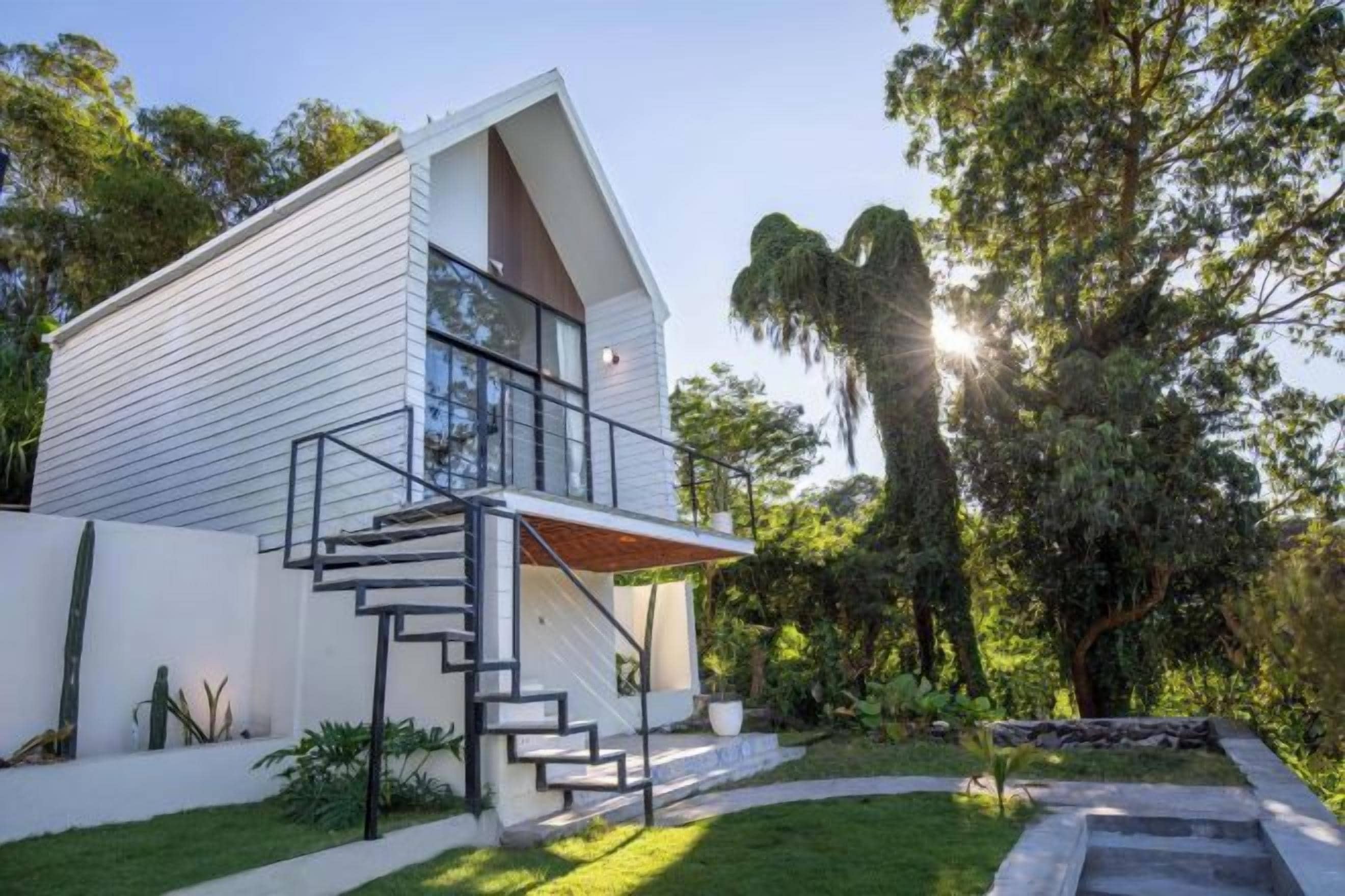 A modern house with a staircase leading to a balcony, surrounded by greenery and trees.