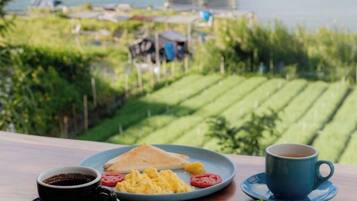 Una mesa con alimentos para desayuno, incluyendo tostadas, huevos y tomates, una taza de café, y vista a un paisaje verde con un cuerpo de agua.