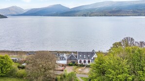 A lakeside property with a large house featuring a prominent chimney, a pathway leading to the house, and a view of the lake and surrounding hills. - Newton Hall (Inveraray)