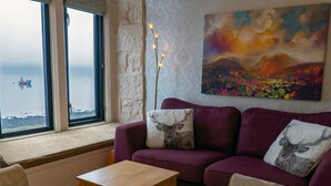 A cozy living room with a purple sofa, two chairs, a small wooden table, and a large window overlooking a body of water. - Newton Hall (Inveraray)