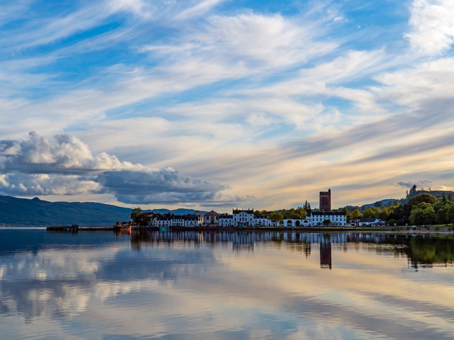 A waterfront town with buildings reflecting on calm water, a prominent tower, and mountains in the background.