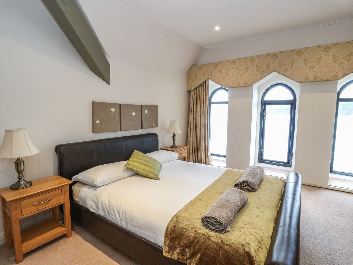 Quarto com uma cama grande, duas mesas de cabeceira, uma janela com cortinas e um painel decorativo na parede.