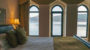 A hotel room with a large bed, a nightstand, a lamp, and three arched windows offering a view of a lake and hills. - Newton Hall (Inveraray)
