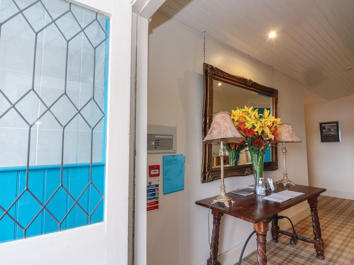A hallway with a blue-tiled wall, a wooden table with a lamp and flowers, a mirror, and a fire action sign.