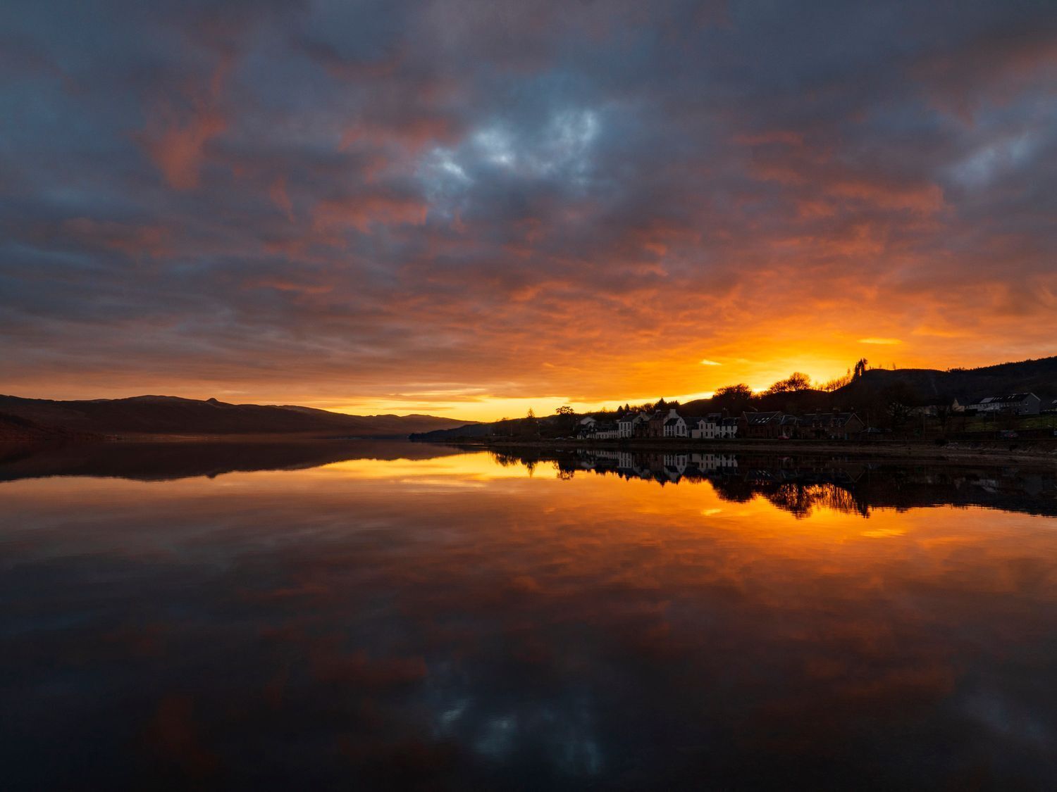 A calm lake reflecting a sunset with a town silhouette across the water.