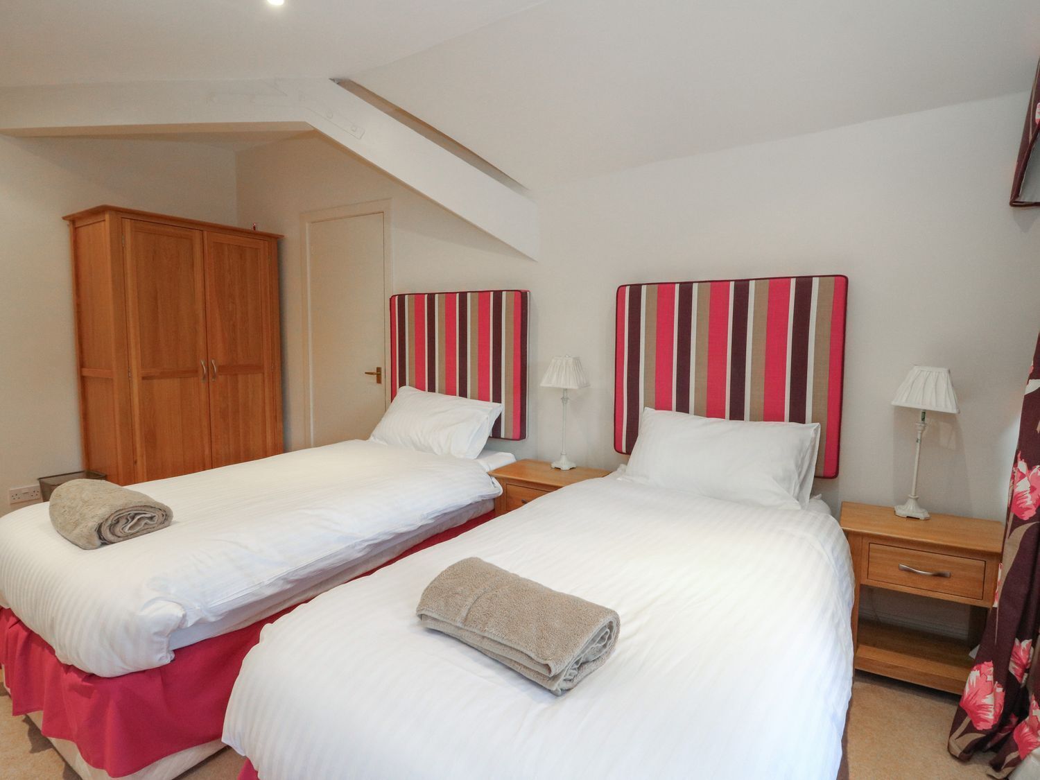 A hotel room with two beds, each with a striped headboard, a wooden wardrobe, and a red bedspread.