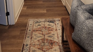 A hallway with wooden flooring, white walls, and a patterned rug. There are doors on both sides, a bench, and a painting on the right wall.