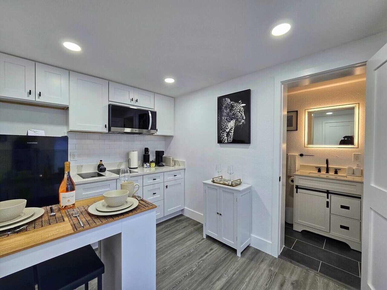 A modern kitchen with white cabinetry, a microwave, a wine bottle, and a dining area with a table set for two.