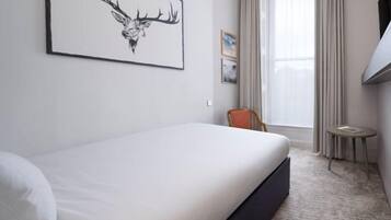 A hotel room with a bed, a chair, a small table, a window with curtains, and a framed picture on the wall.
