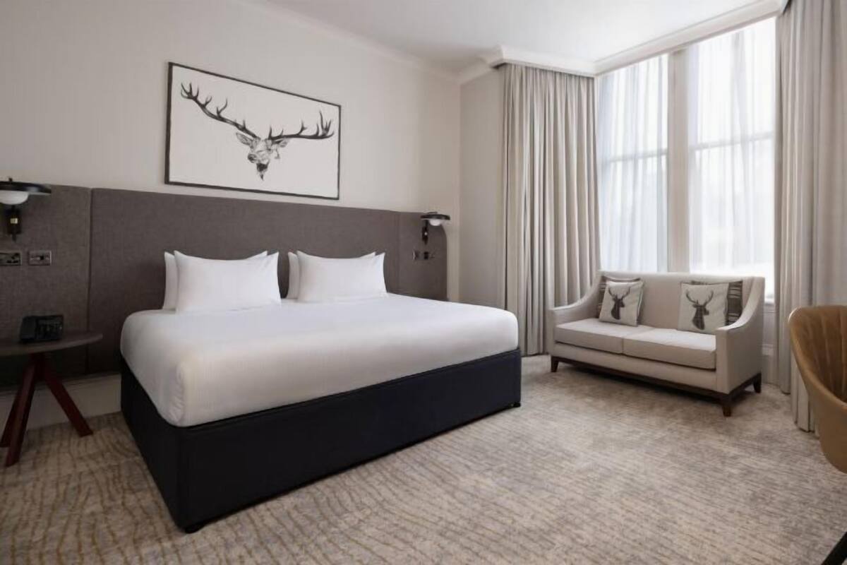 A hotel room with a large bed, a sofa, a desk, and a large window with curtains.
