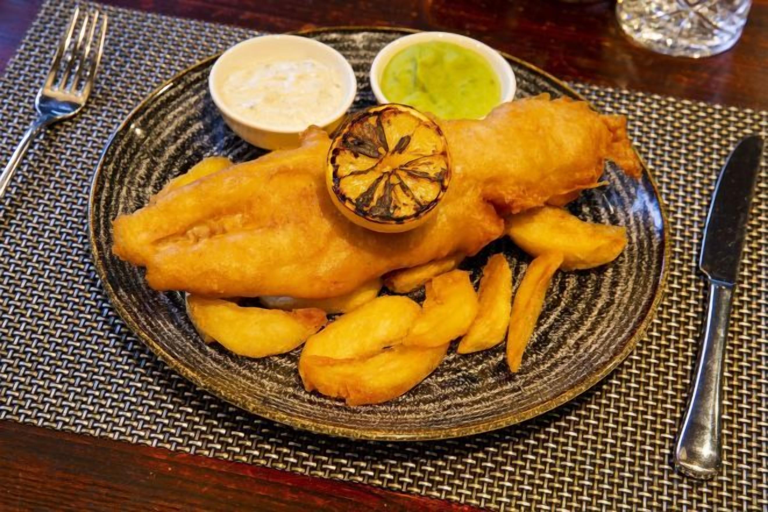Un plato de pescado frito con papas fritas, dos salsas para mojar y un vaso de agua.